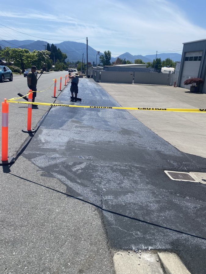 Neill Sealcoating crew member squeegeeing fresh asphalt sealcoat across the Caveman Towing commercial lot in Grants Pass, Oregon