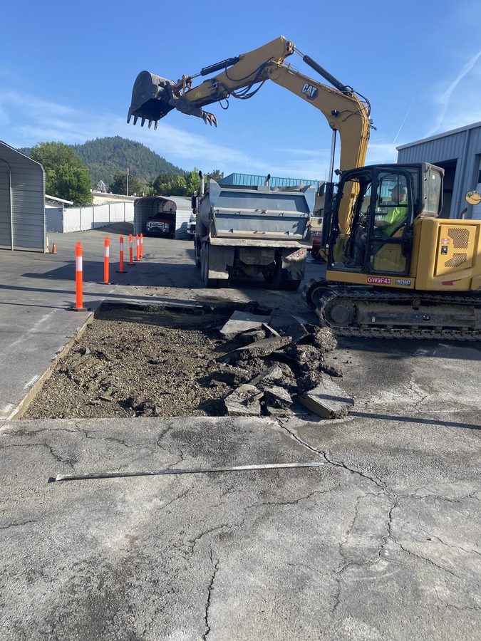 Excavator digging out broken asphalt for a full-depth repair at Caveman Towing in Grants Pass, Oregon
