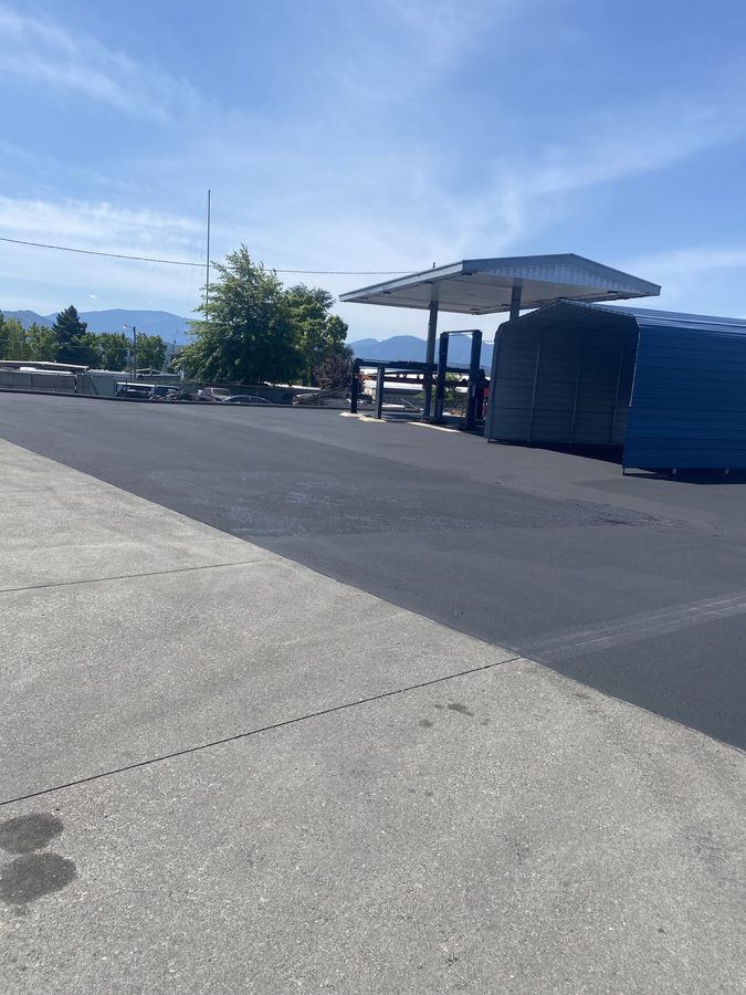 Wide sealcoated commercial lot beside covered equipment bays at Caveman Towing in Grants Pass, Oregon