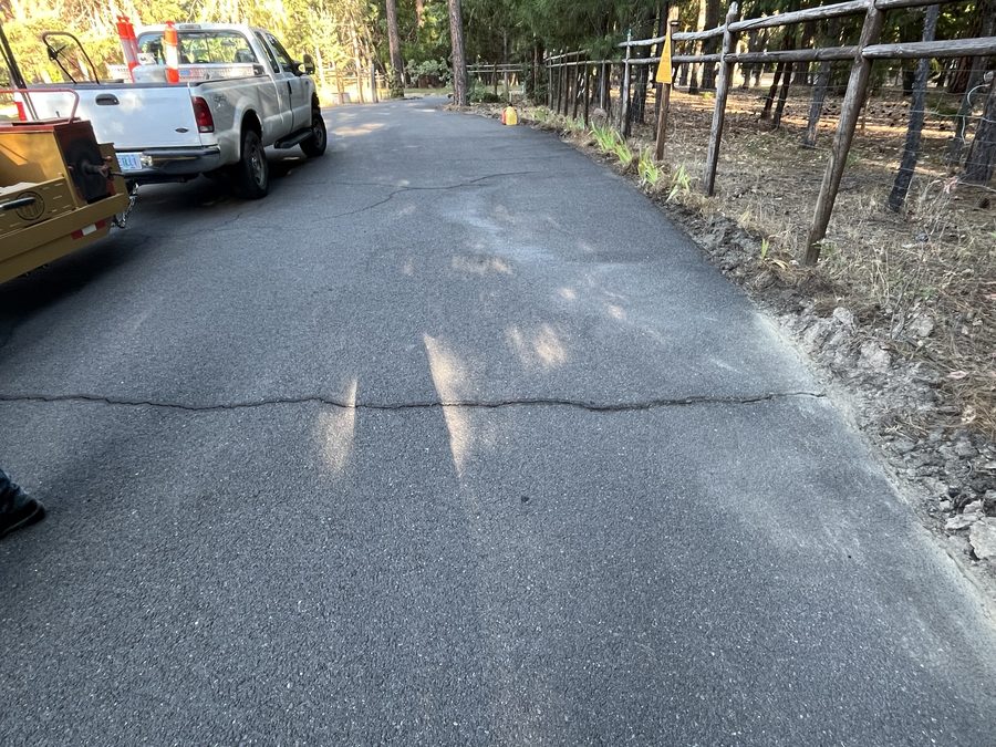 Sun-faded gray asphalt driveway with a long open crack running its length before crack filling on Hidden Valley Road in Grants Pass, Oregon
