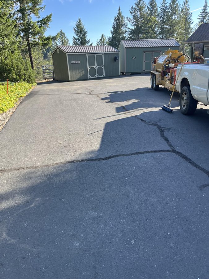 Aging residential asphalt driveway with surface cracks beside two storage sheds before crack filling and sealcoating on Seclusion Loop in Grants Pass, Oregon
