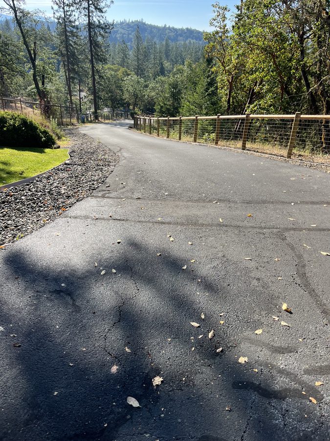 Sun-faded asphalt driveway with spreading surface cracks before crack filling and sealcoating on Seclusion Loop in Grants Pass, Oregon