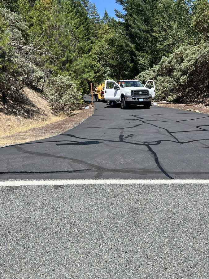 Wide weathered asphalt driveway turnaround with hot-rubber crack fill lines applied before sealcoating on Seclusion Loop in Grants Pass, Oregon