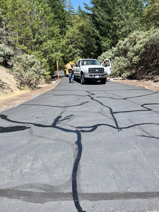 Weathered asphalt driveway approach with crack fill lines and Neill Sealcoating crew visible before sealcoating on Seclusion Loop in Grants Pass, Oregon