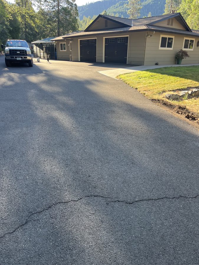 Long crack running across a gray weathered asphalt driveway toward the garage, marked for hot-pour crack filling on Hidden Valley Road in Grants Pass, Oregon
