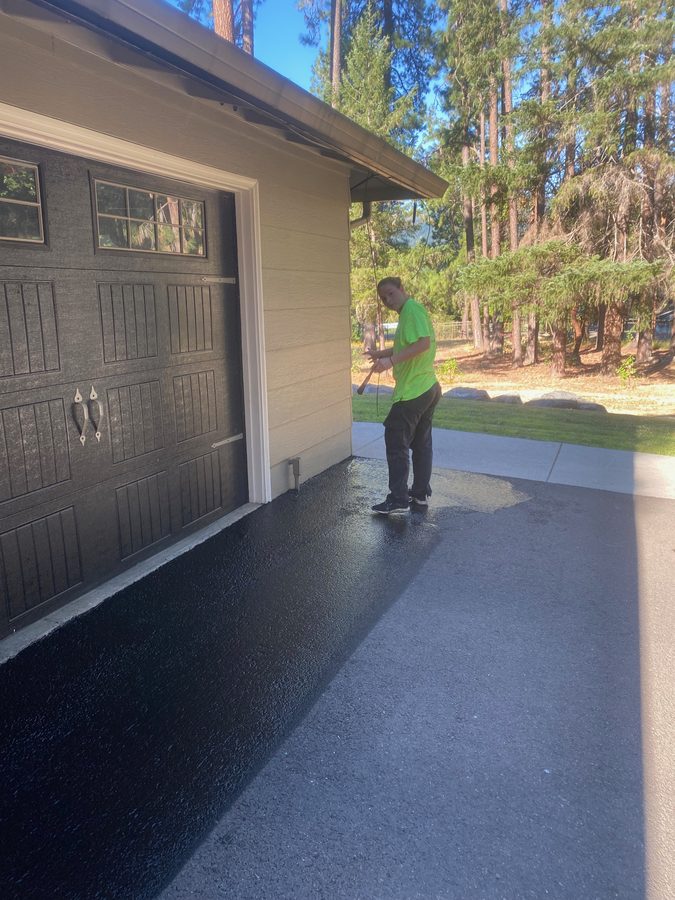 Neill Sealcoating crew member hand-squeegeeing fresh black sealcoat across the driveway apron in front of a garage on Hidden Valley Road in Grants Pass, Oregon
