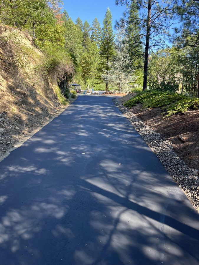 Straight freshly sealcoated residential driveway stretching toward a backyard gazebo lined with pine trees on Seclusion Loop in Grants Pass, Oregon