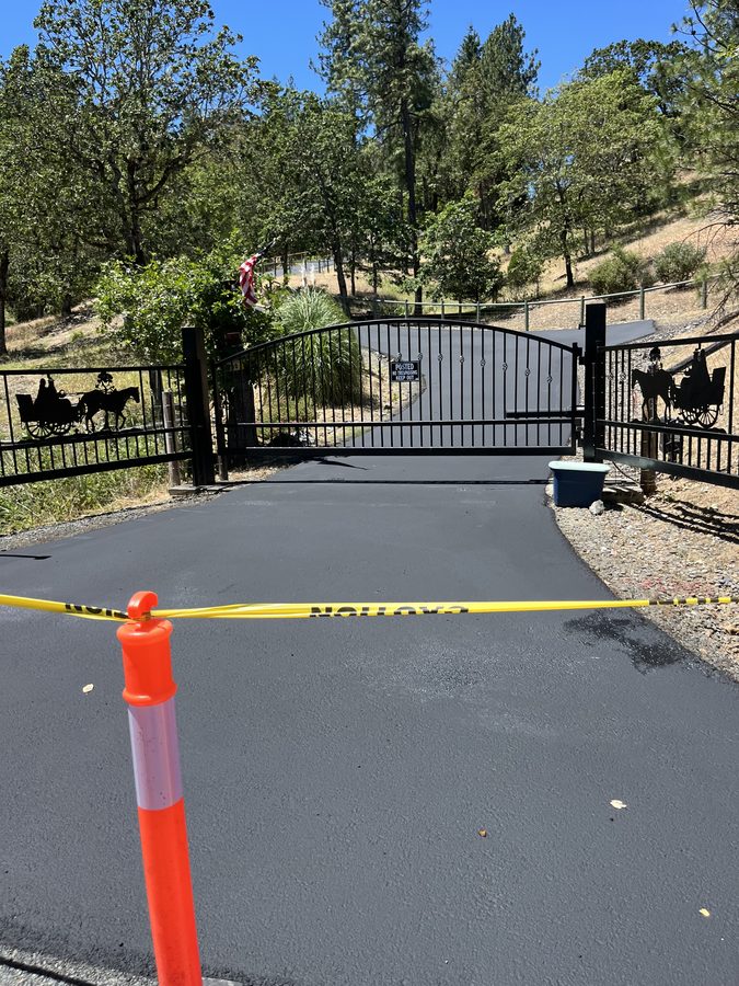 Freshly applied driveway sealcoat blocked off with caution tape and a traffic cone to cure beside a wrought-iron fence on Seclusion Loop in Grants Pass, Oregon