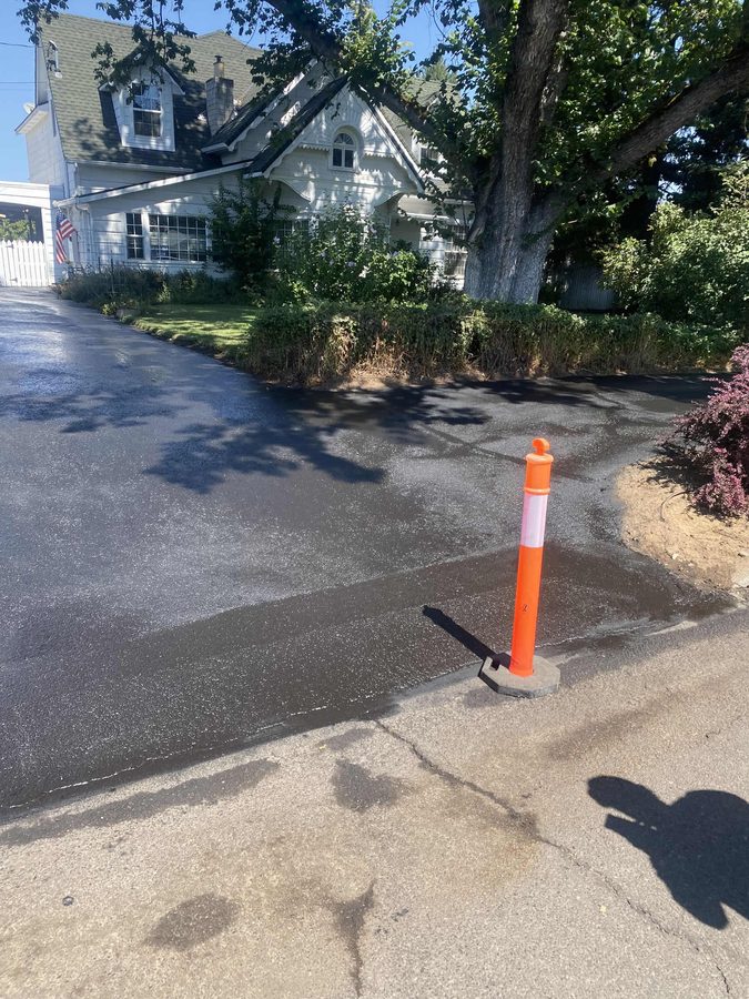Freshly sealcoated residential asphalt driveway curing beside a large shade tree and white farmhouse on Hazel Street in Central Point, Oregon