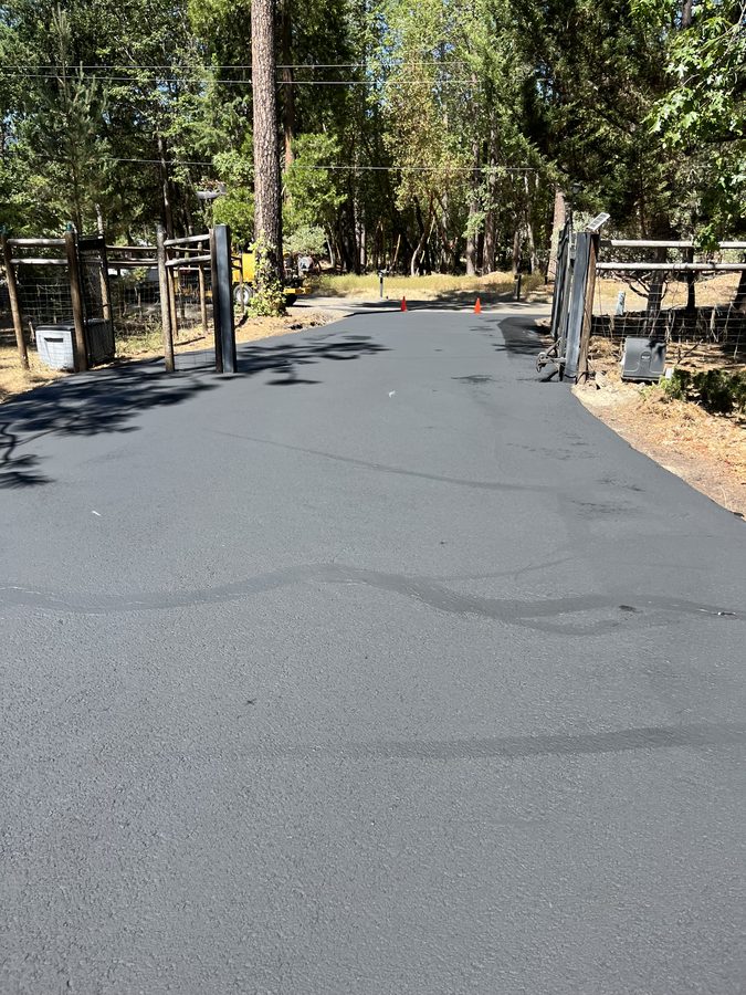 Wide residential asphalt driveway and parking pad freshly sealcoated to a smooth, even finish beside pine trees on Hidden Valley Road in Grants Pass, Oregon