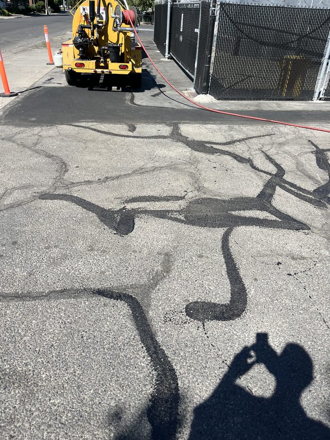 Hot-pour crack-filling kettle on site with fresh rubberized crack fill lines across a worn commercial parking lot on Market Street in Medford, Oregon