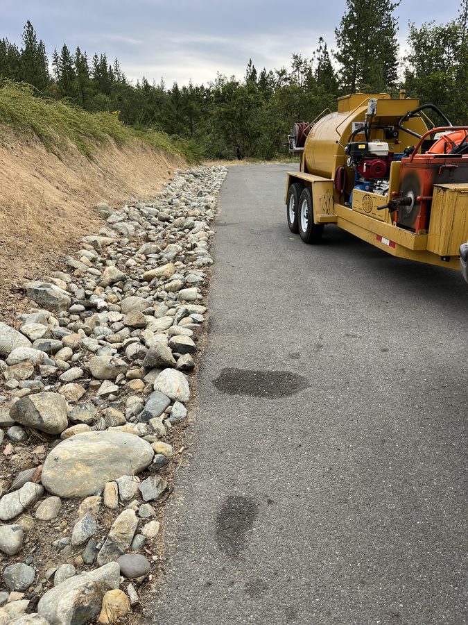 Close-up of a hot-pour crack-filling kettle on a freshly sealcoated driveway beside a river-rock wall on Seclusion Loop in Grants Pass, Oregon