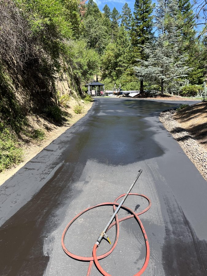 Sealcoat spray wand resting on a freshly applied wet asphalt surface along a hillside private driveway on Seclusion Loop in Grants Pass, Oregon