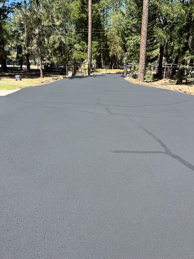 Freshly sealcoated asphalt driveway running clean to the gravel shoulder under tall pines on Hidden Valley Road in Grants Pass, Oregon