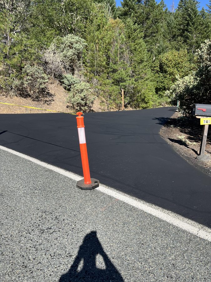 Freshly sealcoated residential driveway entrance with a deep-black finish and an orange traffic cone on Seclusion Loop in Grants Pass, Oregon