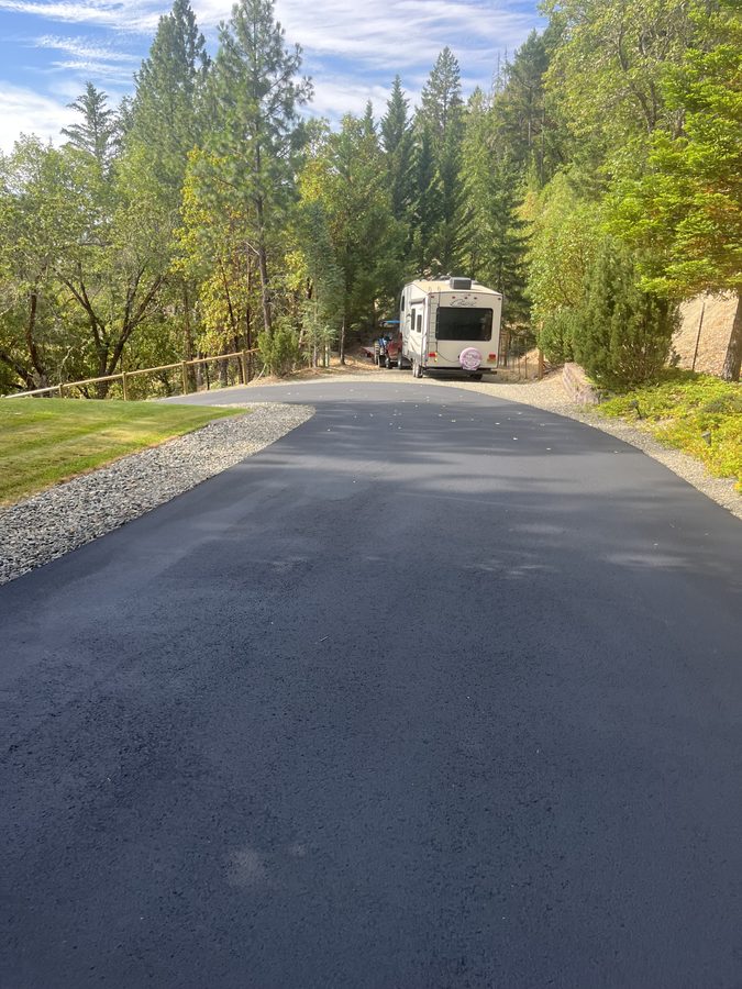 Freshly sealcoated rural asphalt driveway with a gravel shoulder and trees under a clear sky on Seclusion Loop in Grants Pass, Oregon