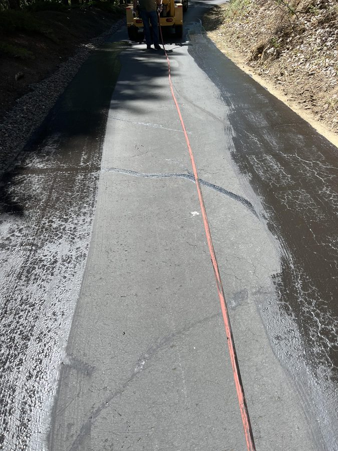 Asphalt driveway sealcoating in progress showing the sealed dark left half and crack-filled gray surface on the right on Seclusion Loop in Grants Pass, Oregon