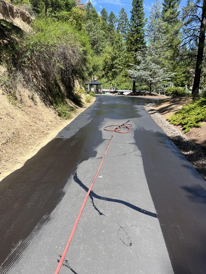 Fresh asphalt sealcoat spray-applied along a long hillside driveway with the sprayer hose still on the wet surface on Seclusion Loop in Grants Pass, Oregon