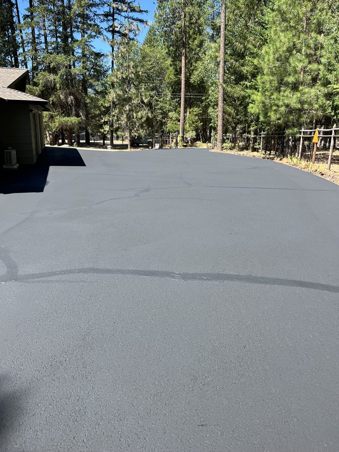 Wide freshly sealcoated asphalt parking pad curving past outbuildings and pine trees on Hidden Valley Road in Grants Pass, Oregon